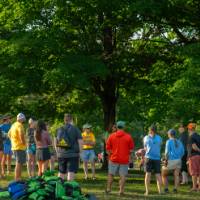 group of adults in shorts and t-shirts standing outside in the sun listening